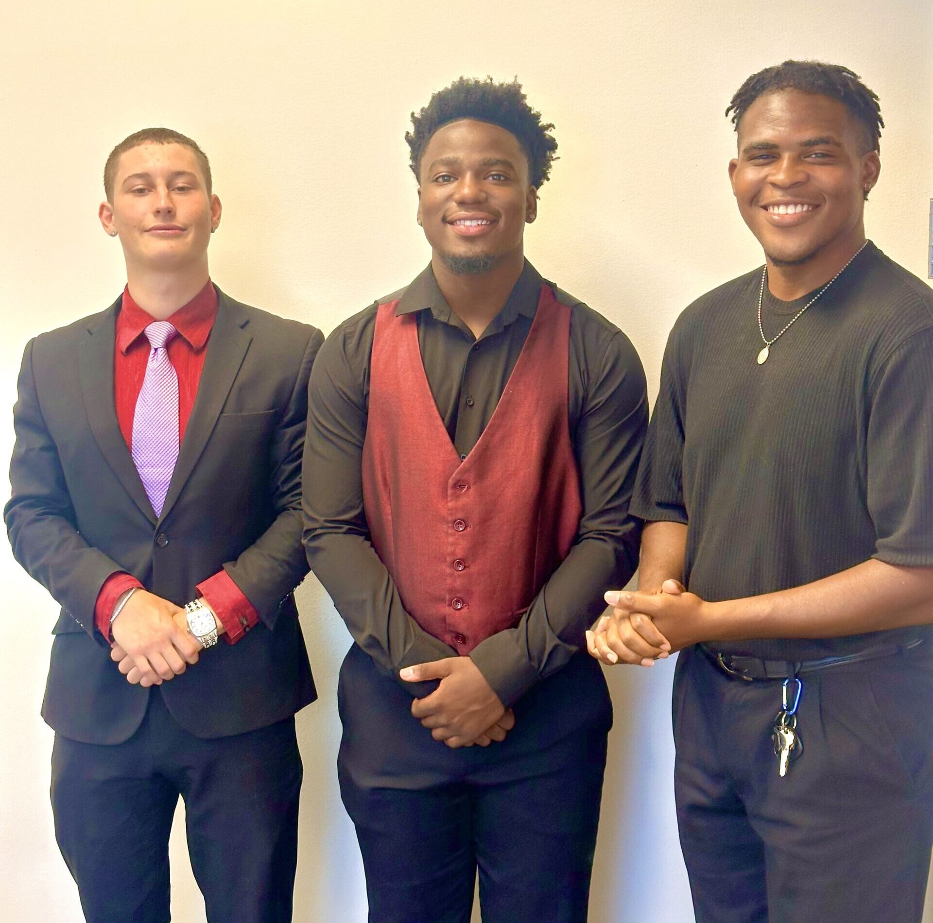 Three professional young men standing together in formal business attire, including a black suit with a red shirt and a maroon vest with a black button-down.